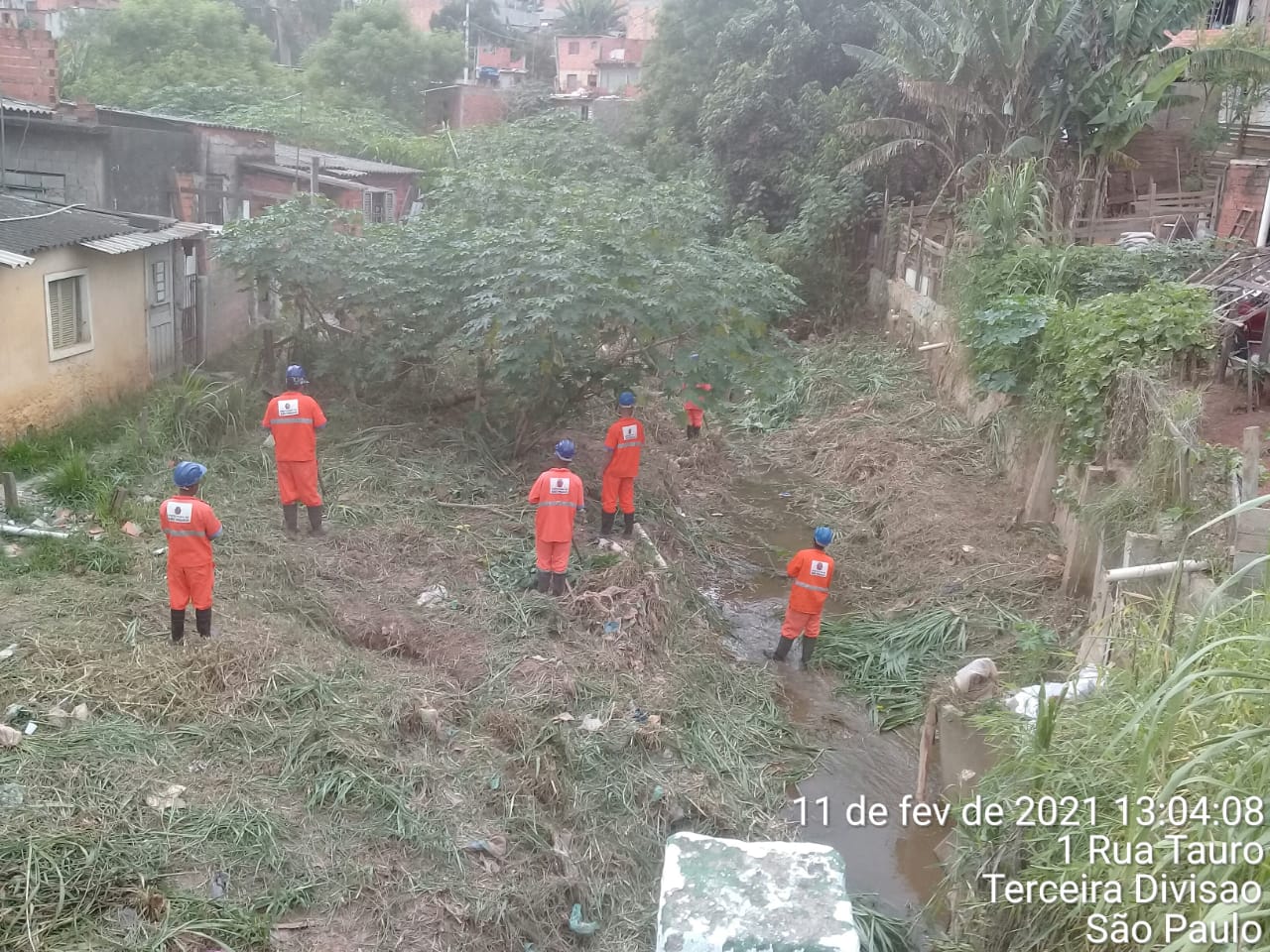 Trabalhadores, com macacão laranja trabalham cortando mato no vale de um córrego 